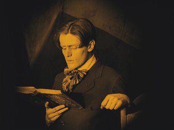 Movie still from “The Cabinet of Dr. Caligari” (1920), directed by Robert Wiene – A man reading a book in a dimly - lit room; Medium shot, Low angle