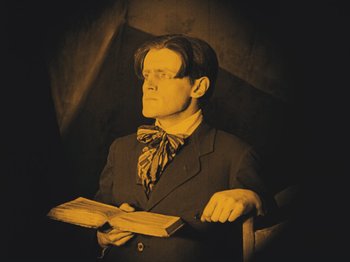 Movie still from “The Cabinet of Dr. Caligari” (1920), directed by Robert Wiene – A man sitting in a chair holding a book in his hands; Medium shot, Low angle