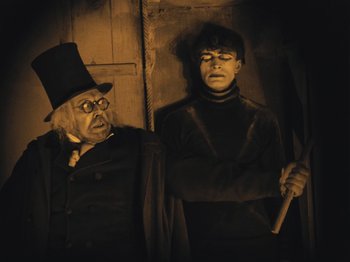 Movie still from “The Cabinet of Dr. Caligari” (1920), directed by Robert Wiene – A man and a woman dressed as a scrooge and the man is holding an axe; Medium shot, Low angle