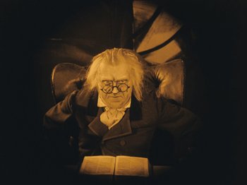 Movie still from “The Cabinet of Dr. Caligari” (1920), directed by Robert Wiene – A man sitting in a chair reading a book; Medium shot, High angle
