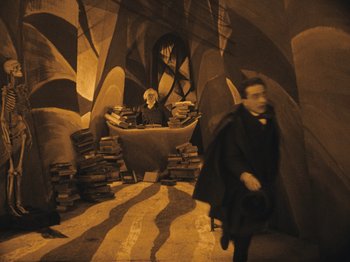 Movie still from “The Cabinet of Dr. Caligari” (1920), directed by Robert Wiene – A man in a black suit and white shirt in a room with books; Wide shot, High angle