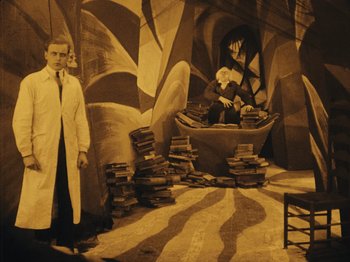 Movie still from “The Cabinet of Dr. Caligari” (1920), directed by Robert Wiene – A man in a lab coat standing in front of stacks of books; Wide shot, Low angle
