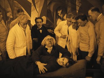 Movie still from “The Cabinet of Dr. Caligari” (1920), directed by Robert Wiene – A group of people standing around a man laying on a bed; Wide shot, High angle