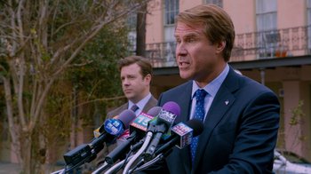Movie still from “The Campaign” (2012), directed by Jay Roach – A man in a suit and tie talking into microphones while another man in a suit looks on; Medium shot, Over the shoulder angle