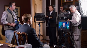 Movie still from “The Campaign” (2012), directed by Jay Roach – A man in a suit is sitting in front of a camera; Medium shot, Over the shoulder angle