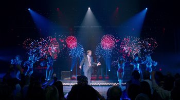 Movie still from “The Campaign” (2012), directed by Jay Roach – A man in a suit is on a stage with fireworks in the background; Wide shot, High angle