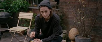 Movie still from “Personal Shopper” (2016), directed by Olivier Assayas – A person sitting on the ground wearing a hat; Medium shot, High angle
