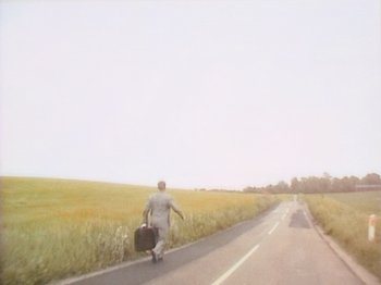 Movie still from “The Celebration” (1998), directed by Thomas Vinterberg – A man walking down the side of a road with a suitcase; Extreme Wide shot, High angle