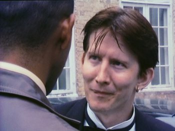 Movie still from “The Celebration” (1998), directed by Thomas Vinterberg – A man in a suit talking to another man in front of a brick building; Close Up shot, Over the shoulder angle