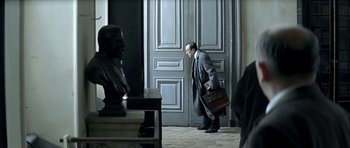Movie still from “The Chorus” (2004), directed by Christophe Barratier – A man with a briefcase walking in front of a statue; Wide shot, Over the shoulder angle