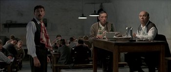 Movie still from “The Chorus” (2004), directed by Christophe Barratier – A man sitting at a wooden table with a laptop; Medium shot, Over the shoulder angle