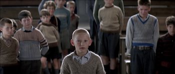 Movie still from “The Chorus” (2004), directed by Christophe Barratier – A young boy with a shaved head stands in front of a group of young boys; Medium shot, Over the shoulder angle