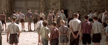 Movie still from “The Chorus” (2004), directed by Christophe Barratier – A group of young men standing around each other on a brick path; Wide shot, High angle