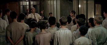 Movie still from “The Chorus” (2004), directed by Christophe Barratier – A group of people are gathered around a man in a room; Medium shot, High angle