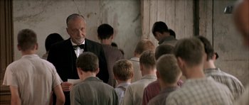 Movie still from “The Chorus” (2004), directed by Christophe Barratier – An old man in a suit and bow tie standing in front of a group of young men; Medium shot, Over the shoulder angle