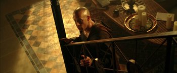 Movie still from “The Chronicles of Riddick” (2004), directed by David Twohy – A bald man leaning on a railing in a room; Medium shot, High angle