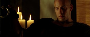 Movie still from “The Chronicles of Riddick” (2004), directed by David Twohy – A man sitting in front of a candle in a dark room; Close Up shot, Over the shoulder angle