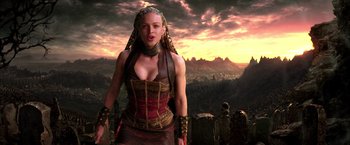 Movie still from “The Chronicles of Riddick” (2004), directed by David Twohy – A woman in a red and brown dress standing in front of mountains; Medium shot, Over the shoulder angle
