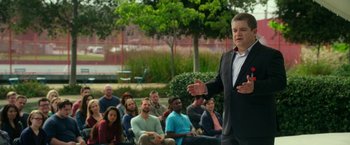 Movie still from “The Circle” (2017), directed by James Ponsoldt – A group of people sitting and standing in front of a man in a business suit; Medium shot, Over the shoulder angle