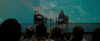 Movie still from “The Circle” (2017), directed by James Ponsoldt – A man and a woman sitting on chairs in front of an audience; Wide shot, Over the shoulder angle