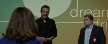 Movie still from “The Circle” (2017), directed by James Ponsoldt – A man standing in front of a woman in front of a projection screen; Close Up shot, Over the shoulder angle