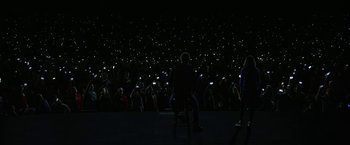 Movie still from “The Circle” (2017), directed by James Ponsoldt – A group of people sitting in front of a crowd at night with cell phones; Extreme Wide shot, High angle