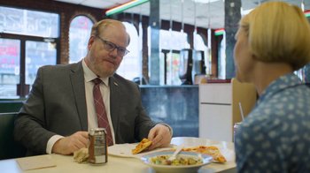 Movie still from “Full Circle” (2023), directed by Steven Soderbergh – A man sitting at a table with a plate of pizza; Medium shot, Over the shoulder angle
