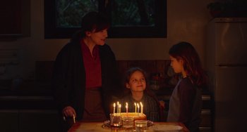 Movie still from “Petite Maman” (2021), directed by Céline Sciamma – A woman and two children standing in front of lit candles on a cake; Medium shot, Over the shoulder angle