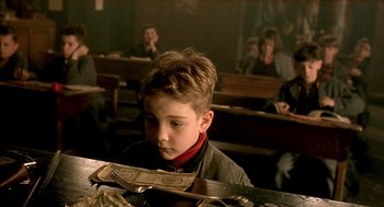 Movie still from “The City of Lost Children” (1995), directed by Jean-Pierre Jeunet – A young boy sitting at a table in front of a group of people; Close Up shot, Over the shoulder angle