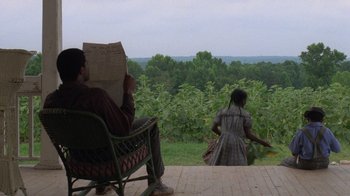 Movie still from “The Color Purple” (1985), directed by Steven Spielberg – A man sitting in a rocking chair reading a newspaper; Wide shot, Over the shoulder angle