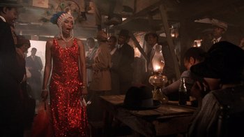 Movie still from “The Color Purple” (1985), directed by Steven Spielberg – A group of people standing around a table with a red dress on; Medium shot, Over the shoulder angle