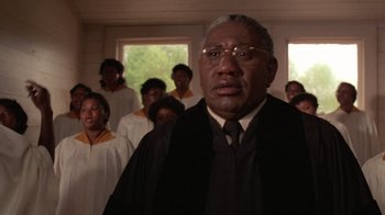 Movie still from “The Color Purple” (1985), directed by Steven Spielberg – An older man in a robe and glasses stands in front of a group of people; Close Up shot, Low angle