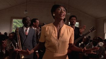 Movie still from “The Color Purple” (1985), directed by Steven Spielberg – A woman in a yellow dress singing in front of a group of men; Medium shot, Low angle