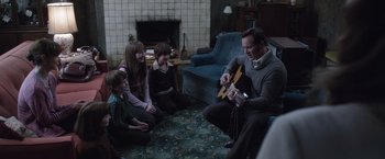 Movie still from “The Conjuring 2” (2016), directed by James Wan – A group of people sitting in a living room with a man playing a guitar; Medium shot, High angle