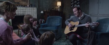 Movie still from “The Conjuring 2” (2016), directed by James Wan – A group of people sitting in a living room with a man playing an acoustic guitar; Medium shot, Over the shoulder angle