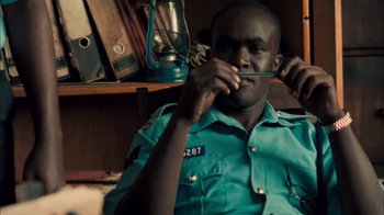 Movie still from “The Constant Gardener” (2005), directed by Fernando Meirelles – A man sitting in front of a bookshelf holding a knife to his mouth; Medium shot, Over the shoulder angle
