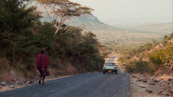 Movie still from “The Constant Gardener” (2005), directed by Fernando Meirelles – A person walking down a road with a car on the side of the road; Extreme Wide shot, Low angle