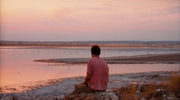 Movie still from “The Constant Gardener” (2005), directed by Fernando Meirelles – A man sitting on top of a rock looking out at a body of water; Wide shot, Over the shoulder angle