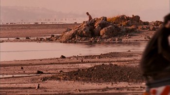 Movie still from “The Constant Gardener” (2005), directed by Fernando Meirelles – A man sitting on a rock on the beach; Extreme Wide shot, High angle