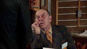 Movie still from “The Conversation” (1974), directed by Francis Ford Coppola – An older man wearing a suit and tie while sitting at a table; Medium shot, Over the shoulder angle