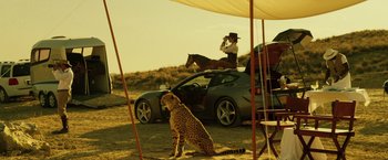 Movie still from “The Counselor” (2013), directed by Ridley Scott – A cheetah sitting on the ground next to a parked car; Wide shot, Low angle