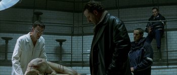 Movie still from “The Crimson Rivers” (2000), directed by Mathieu Kassovitz – A man standing next to a table in a room; Medium shot, Over the shoulder angle