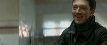 Movie still from “The Crimson Rivers” (2000), directed by Mathieu Kassovitz – A man wearing a black leather jacket smiling for the camera; Close Up shot, Over the shoulder angle