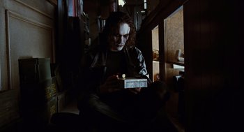 Movie still from “The Crow” (1994), directed by Alex Proyas – A man sitting in a room holding a box; Medium shot, Low angle