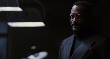 Movie still from “The Crow” (1994), directed by Alex Proyas – A man in a suit and glasses is standing in a dark room; Close Up shot, Over the shoulder angle