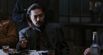 Movie still from “The Crow” (1994), directed by Alex Proyas – A man sitting at a table with a cigarette in his mouth; Medium shot, Over the shoulder angle