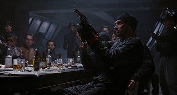 Movie still from “The Crow” (1994), directed by Alex Proyas – A man sitting at a table holding a gun in his hand; Medium shot, Over the shoulder angle