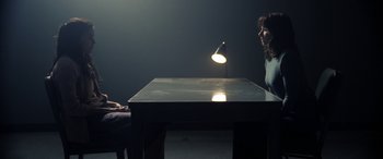 Movie still from “The Curse of La Llorona” (2019), directed by Michael Chaves – A woman sitting at a table in a dark room; Medium shot, Over the shoulder angle