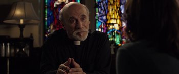 Movie still from “The Curse of La Llorona” (2019), directed by Michael Chaves – An older man wearing a priest's outfit in front of a stained glass window; Close Up shot, Over the shoulder angle