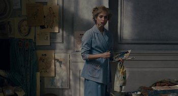 Movie still from “The Danish Girl” (2015), directed by Tom Hooper – A woman standing in front of a wall holding a painting; Medium shot, Over the shoulder angle
